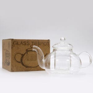 A clear glass teapot with a lid is placed next to a brown cardboard box labeled "GLASS TEAPOT" on a plain white background.