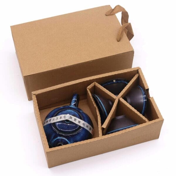 A cardboard box containing a blue ceramic teapot and two teacups, separated by cardboard dividers, with the box lid and handles visible.