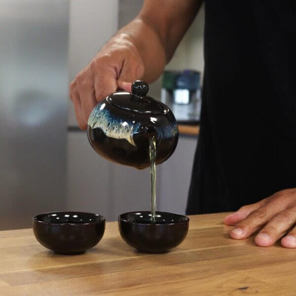 A person pours tea from a ceramic teapot into one of two black teacups on a wooden table.