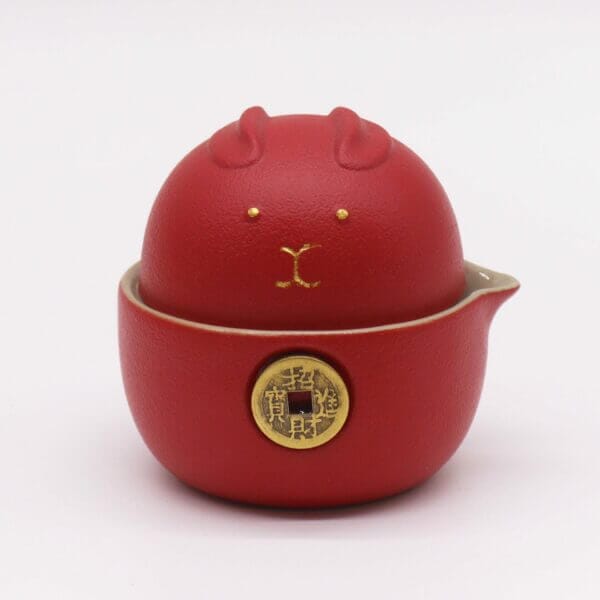 A red ceramic container with a lid shaped like a stylized animal face, featuring gold accents and a gold circular emblem on the front.