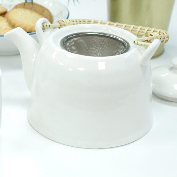 White ceramic teapot with a bamboo handle and a metal strainer inside, placed on a white surface with a plate of cookies in the background.