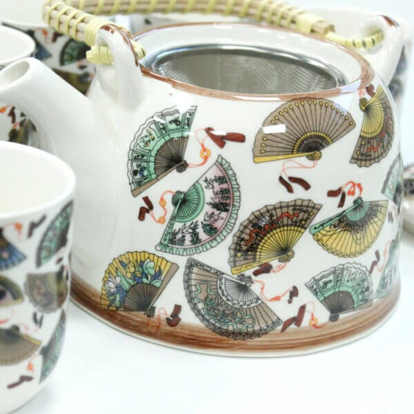White ceramic teapot decorated with colorful hand fan patterns, featuring a woven handle and accompanied by matching teacups.