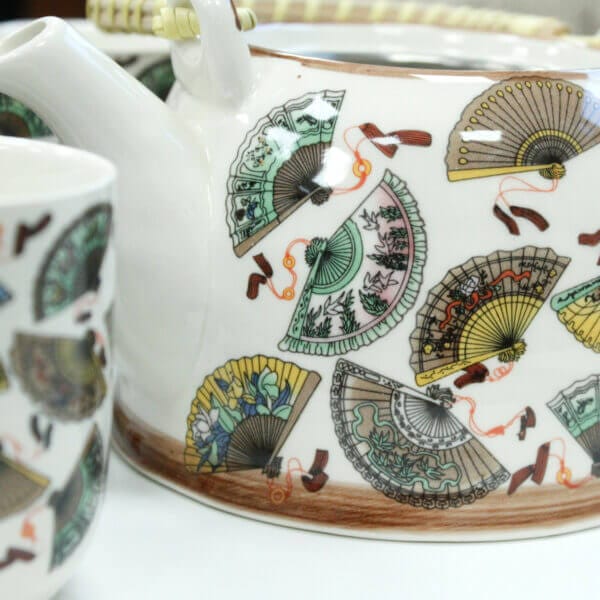 Close-up of a ceramic teapot and cup decorated with colorful hand fan patterns and a bamboo handle.