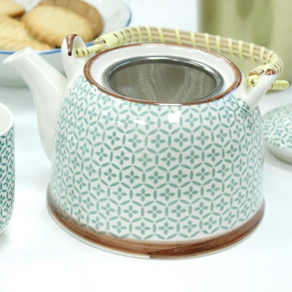 A patterned ceramic teapot with a metal strainer inside, featuring a handle wrapped in yellow material. A plate of cookies and a cup are partially visible in the background.