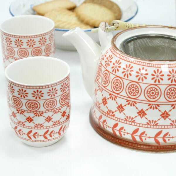A white teapot and two matching cups with red patterns are placed next to a plate of rectangular biscuits.