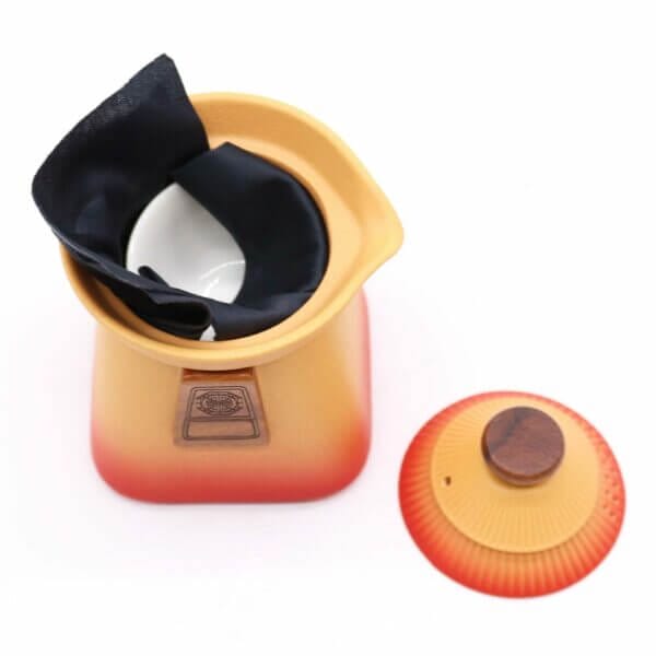 A ceramic container with a red-yellow gradient, containing a white bowl and black cloth, sits next to its matching lid with a wooden knob.