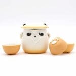 A tea set featuring a panda-shaped teapot with a lid and two matching cups, all with a beige and white color scheme and paw print details.