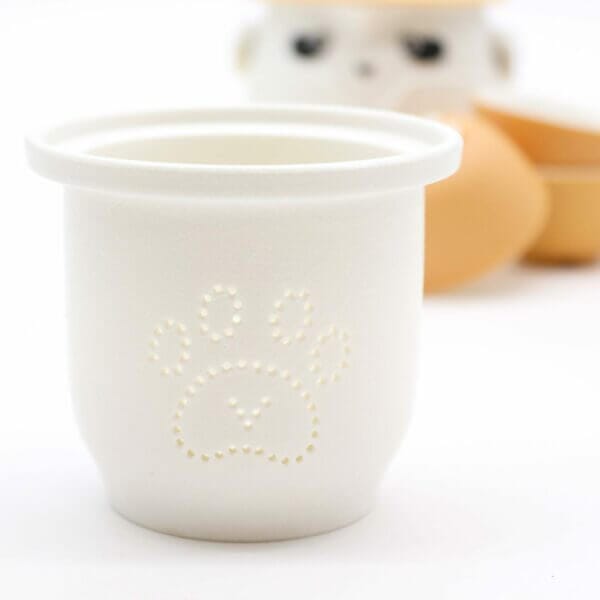 A white cup with a paw print design on the front sits in focus; blurred animal-shaped objects and eggs are visible in the background.