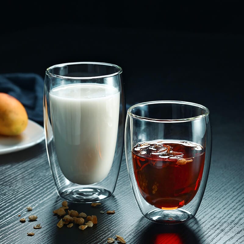 Two Oval-Shape Double-Wall Glass Cups rest on a table—one with milk and the other with iced tea. A partially visible plate holding a pear enhances the elegance of this serene scene.