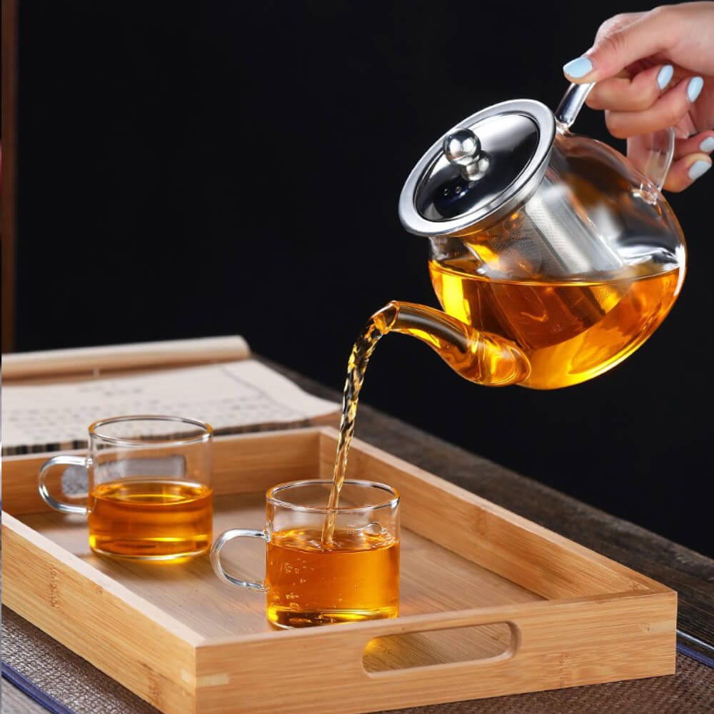 A hand pours tea from the Elegant Glass Teapot with Stainless Steel Infuser (800ml) into a cup, while another cup rests on a wooden tray next to a closed book.